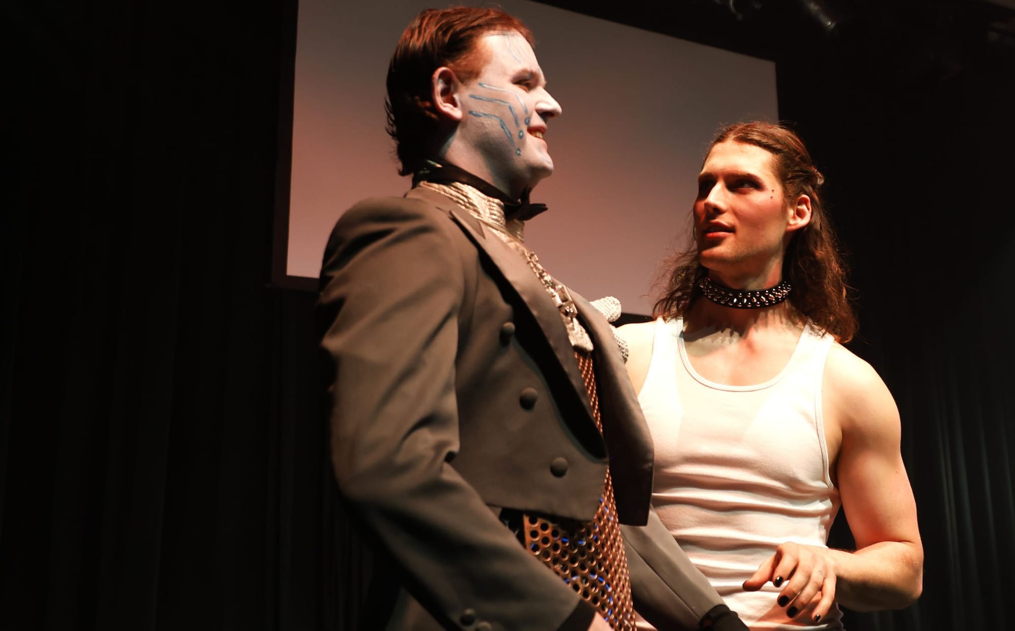 Photo of a man in a robot costume with make-up and a muscly man in a white singlet and studded collar.