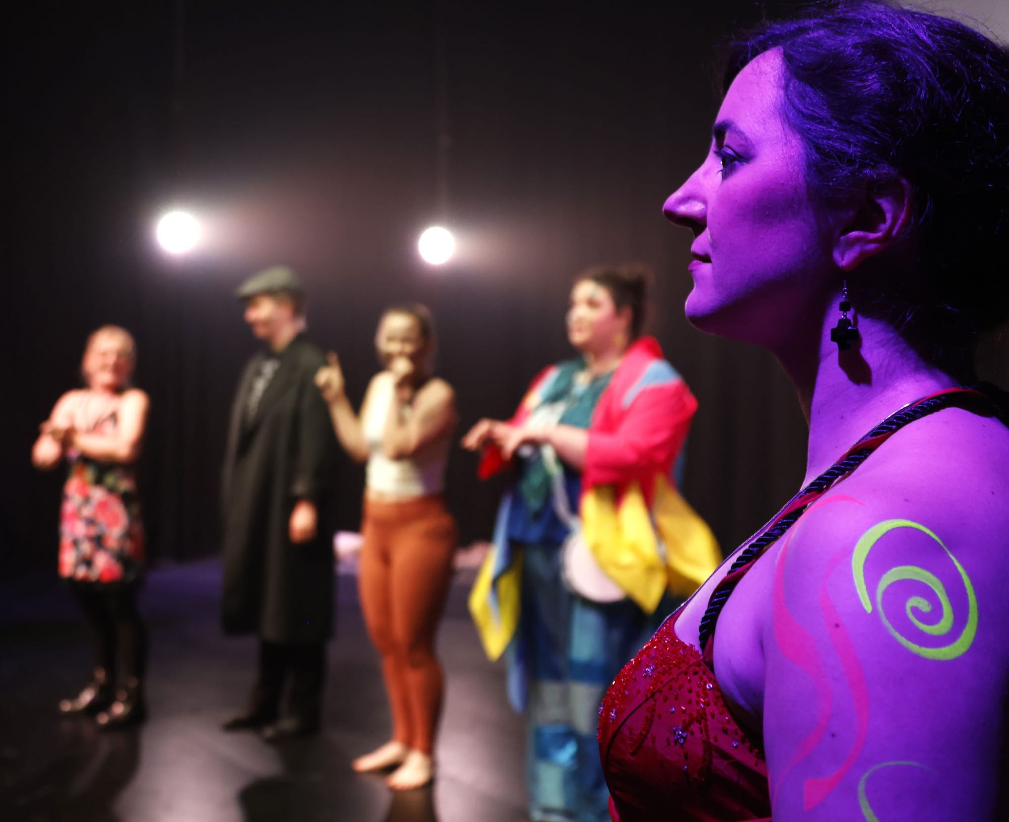A colourful line-up of mostly-out-of-focus people. Bernadette Richardson as Goneril in the foreground is lit with purple and has fluorescent swirls on her bare arm.