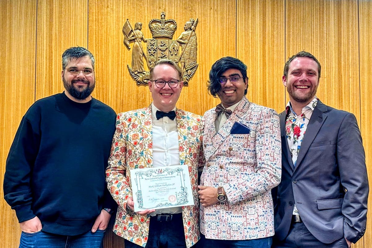 Three people pose beneath the New Zealand Coat of Arms. Some are dressed flamboyantly. Mark is holding a certificate.