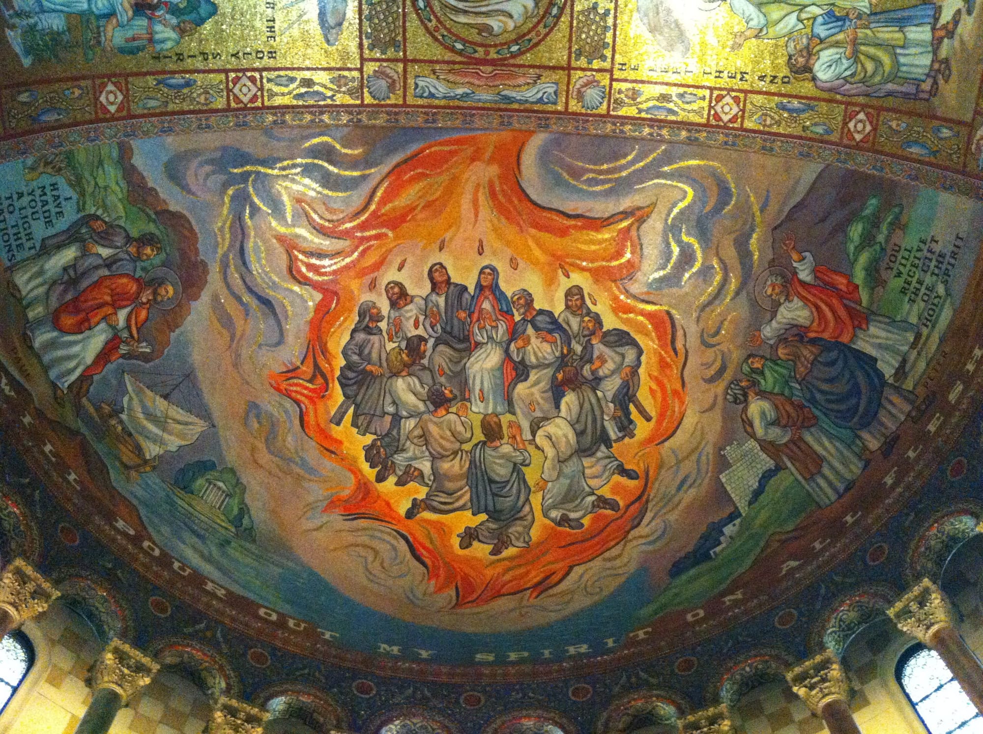 A mosaic on a cathedral ceiling depicts the descending fire of the Holy Spirit on the Day of Pentecost, with a flame floating above each person’s head. Text around the mosaic says "I have made you a light to the nations”, “I will pour out my Spirit on all flesh”, and “You will receive the gift of the Holy Spirit".