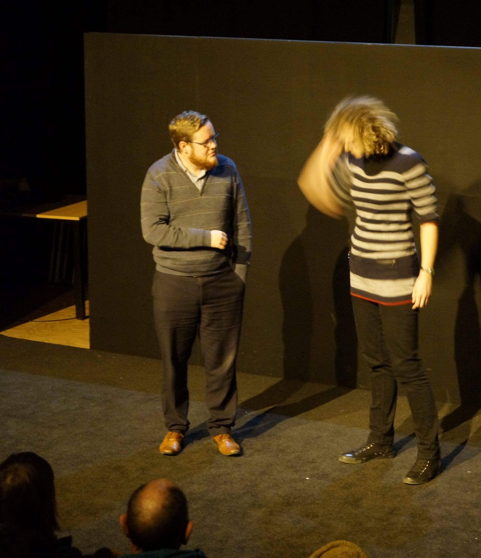 Mark performs with a friend on a black stage. Both are wearing monochrome clothes. He has a beard and a concerned pose. She has frizzy hair. Her arm and head are blurry due to motion.