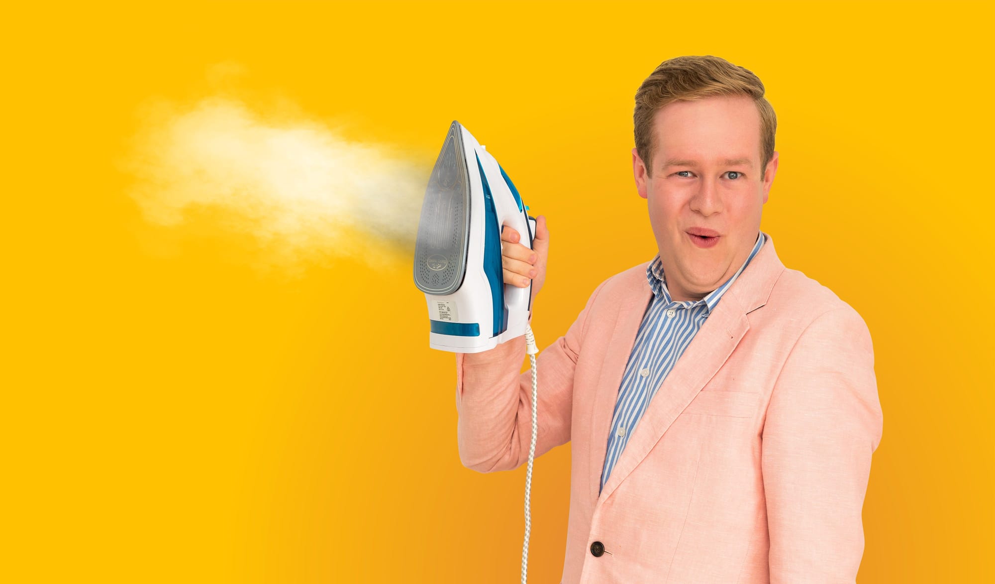 Promotional image with a deep yellow background. Mark wears a baby pink suit and wields a steaming iron. He has a salacious facial expression.