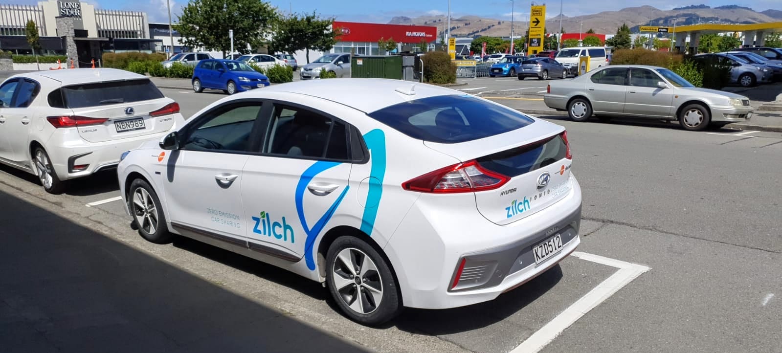 Photo of a white Zilch car parked on a street with Lone Star, Pak'nSave, and the Port Hills in the background.