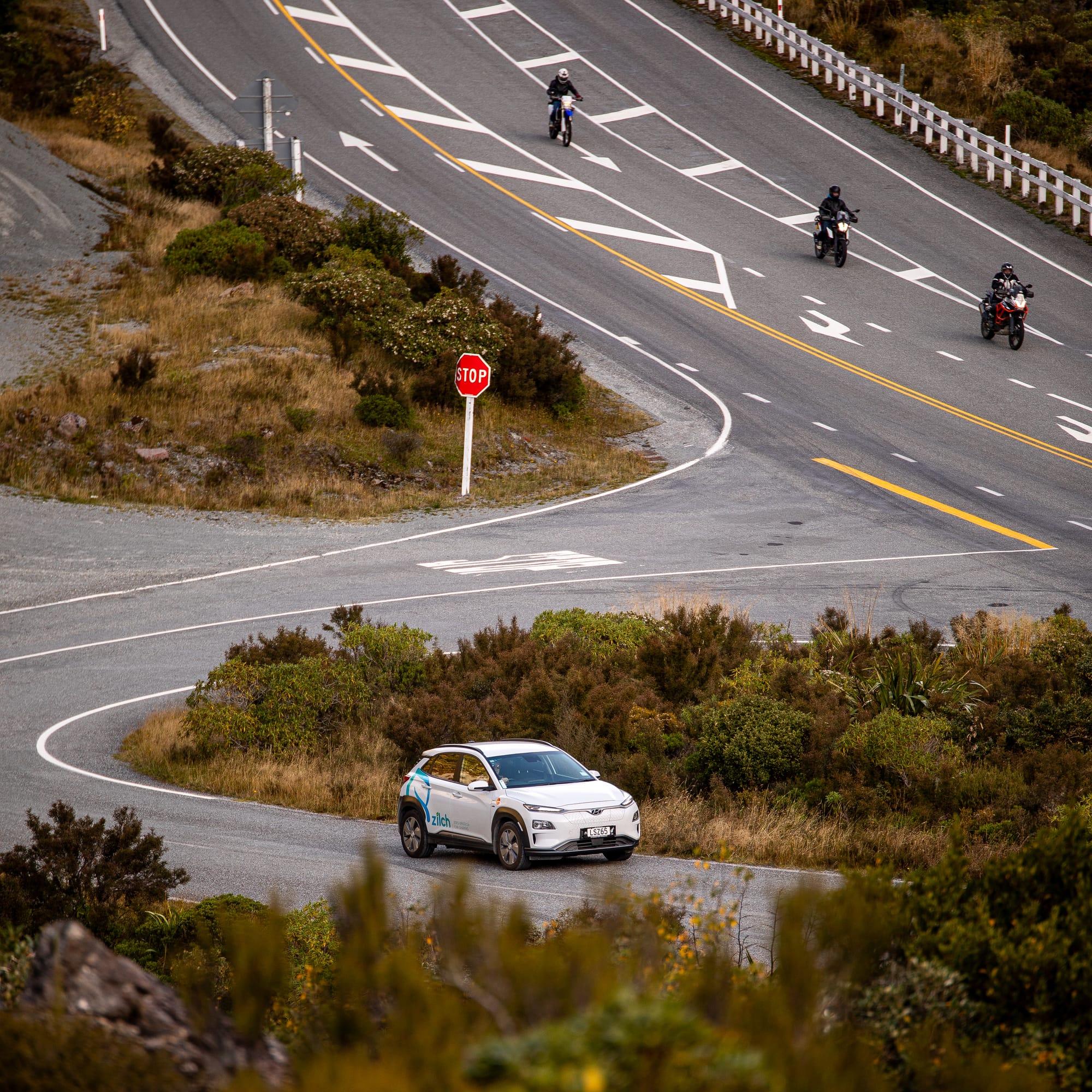 Photo of a white Hyundai Kona (with blue Zilch logo) driving uphill from an intersection. There is parched vegetation on both sides of the road. In the background are three motorcyclists.