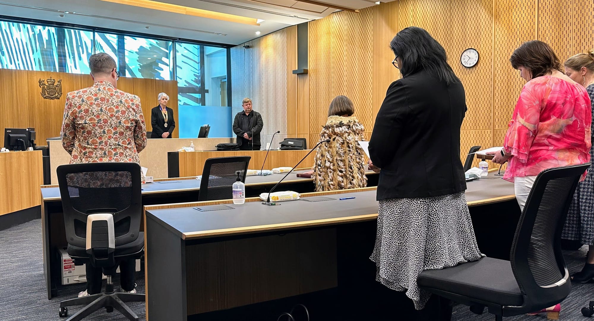 People standing in a courtroom. The Justices being sworn in are facing away from the camera. One has her hand on a Bible. Mark is on the left in his floral blazer.